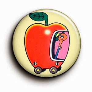 Puede incluir: Un botón redondo con un diseño de manzana de dibujos animados. La manzana es roja con una hoja verde y un conductor en un coche rosa. El fondo es de color amarillo claro.