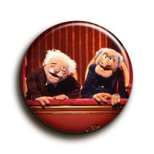 Könnte beinhalten: Ein runder Button mit Statler und Waldorf, den beiden mürrischen Muppet Show-Charakteren, in einer Theaterloge. Der Hintergrund ist rot, und die Puppen sind im Vordergrund, mit ihren Händen auf der Loge.