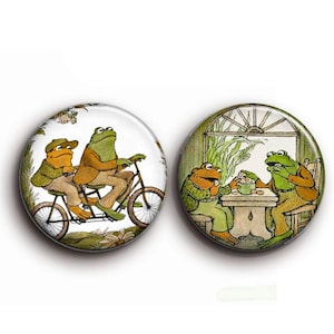 Könnte beinhalten: Zwei runde Buttons mit Illustrationen von anthropomorphen Fröschen. Einer zeigt zwei Frösche auf einem Tandem, der andere zeigt zwei Frösche an einem Tisch. Die Buttons haben einen weißen Hintergrund.