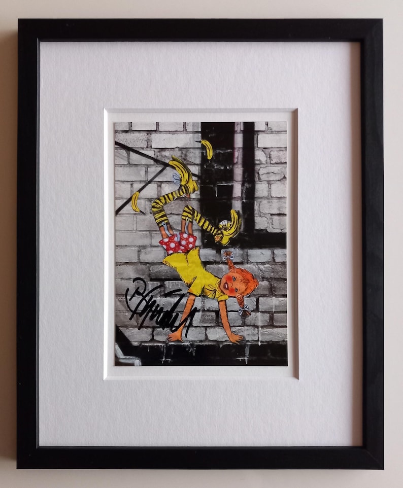 Puede incluir: Una ilustraci&oacute;n enmarcada de una ni&ntilde;a haciendo una parada de manos contra una pared de ladrillo. Lleva un vestido amarillo, medias de lunares rojos y blancos y calcetines a rayas amarillas y negras. Hay pl&aacute;tanos amarillos cayendo a su alrededor. La ilustraci&oacute;n est&aacute; firmada "Brenda Z" en la esquina inferior izquierda.