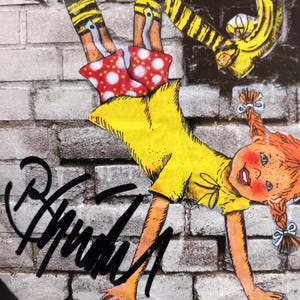 Puede incluir: Una ni&ntilde;a de dibujos animados amarilla, roja y negra haciendo una parada de manos en una pared de ladrillo. La ni&ntilde;a lleva pantalones amarillos con rayas negras, una camisa amarilla y una falda roja con lunares blancos. La ni&ntilde;a tiene el pelo rojo y una gran sonrisa. La imagen est&aacute; firmada "Anarchy" con un marcador negro.