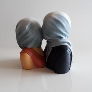 Könnte beinhalten: Keramikskulptur, die zwei Figuren in einer Umarmung darstellt. Eine Figur trägt ein rotes Kleid, die andere einen schwarzen Anzug. Beide Figuren haben ihre Gesichter mit einem hellblauen Tuch bedeckt, was eine mysteriöse und künstlerische Ästhetik erzeugt.