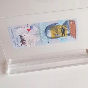 Peut inclure: Un présentoir en acrylique transparent avec un billet de 20 bolivars représentant un portrait de Simon Bolivar et un cœur rouge avec un ballon bleu. Le billet porte le texte "Republica Bolivariana de Venezuela" et le numéro de série "B78303772".