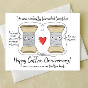 May include: A white greeting card with a cotton anniversary theme. The card features two cartoon spools of thread, a red heart, and the text "We are perfectly threaded together." Additional text includes "Happy Cotton Anniversary!" and "2 amazing years ago, we tied the knot."