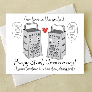 May include: A white greeting card with a humorous design for a steel anniversary. The card features two cartoon graters, a red heart, and the text "Our Love is the gratest" and "Happy Steel Anniversary!"