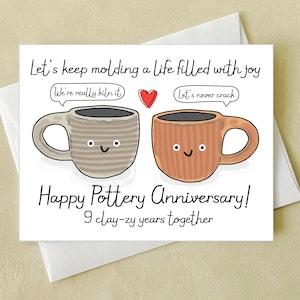May include: A greeting card with the text "Let's keep molding a life filled with joy" and "Happy Pottery Anniversary!" features two cartoon mugs with faces and a red heart. The card is on a white envelope.