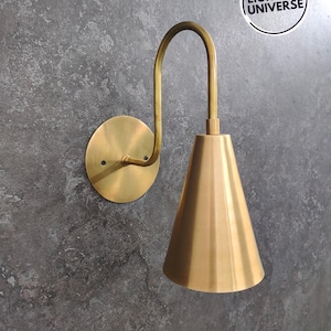 Peut inclure: Une applique murale dorée avec un bras incurvé et un abat-jour conique. L'applique est montée sur un mur gris.