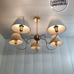 May include: A gold chandelier with five light bulbs and white fabric lampshades. The chandelier is hanging from a white ceiling.