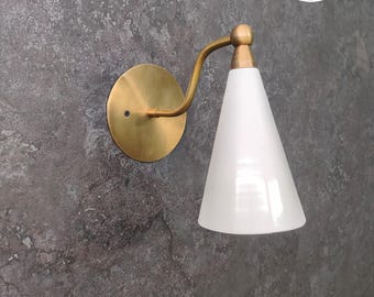 May include: A brass wall sconce with a white cone-shaped shade. The sconce has a simple, modern design and is perfect for adding a touch of elegance to any room.