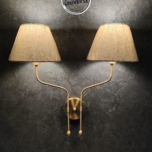 May include: A gold wall sconce with two beige lampshades. The sconce has a curved design and is mounted on a dark grey wall.