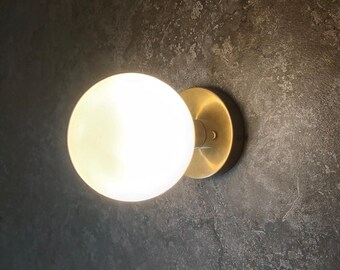May include: A round, white globe light fixture with a brass-colored base. The light is illuminated, casting a warm glow. The fixture is mounted on a dark, textured wall, creating a modern aesthetic.