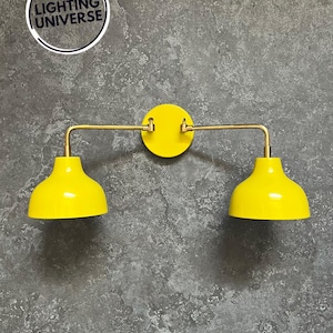 Op de afbeelding: Een paar gele wandlampen met gouden metalen armen. De lampen hebben een eenvoudig, modern ontwerp en zijn perfect om elke kamer een vleugje kleur te geven.