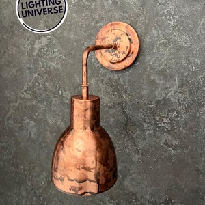 May include: A copper wall sconce with a distressed finish. The sconce has a round back plate and a curved arm that supports a bell-shaped shade.