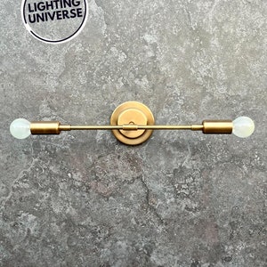 May include: A gold metal wall sconce with two light bulbs. The sconce has a simple, modern design with a straight bar and two round bulbs.