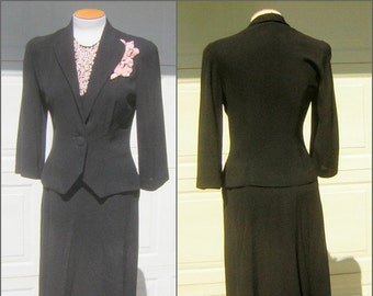 May include: A black skirt suit with a fitted jacket and a skirt that falls to the knee. The jacket has a single button closure and a small pink flower brooch on the lapel.