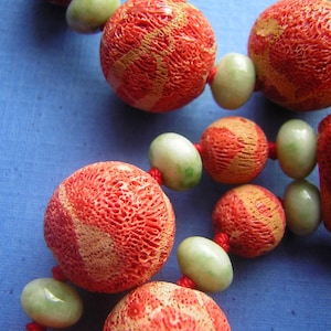 K&ouml;nnte beinhalten: Nahaufnahme einer roten und orangefarbenen Korallenperlenkette. Die Perlen sind rund und haben eine strukturierte Oberfl&auml;che. Die Kette ist auf einer roten Schnur aufgef&auml;delt.