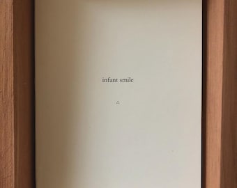 Könnte beinhalten: Ein weißes Blatt Papier mit dem Text "infant smile" in schwarzer Schrift. Das Papier befindet sich in einem Holzrahmen. Unter dem Text befinden sich zwei kleine Punkte. Der Rahmen hat eine hellbraune Oberfläche.