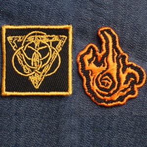 May include: Two embroidered patches, one with a gold triangle design on a black background and the other with an orange flame design on a black background.