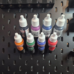 May include: A collection of eight small bottles of acrylic paint. The bottles are arranged in a row on a black surface. The bottles are labelled with different colours, including orange, green, blue, red, black, purple, and dark blue. The bottles are all the same size and shape, and they have a white cap.