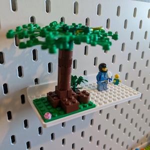 May include: A white pegboard with a small white shelf attached. The shelf has a green and brown Lego tree, a blue Lego minifigure, and a yellow Lego minifigure.