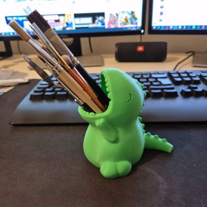 May include: A green dinosaur-shaped pencil holder with a wide open mouth, holding a variety of paintbrushes and art tools.