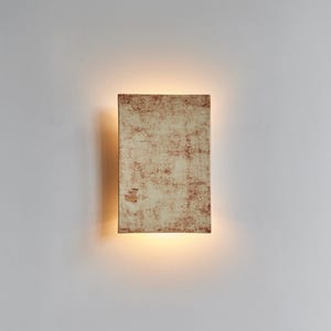 Può includere: Applique da parete rettangolare con superficie strutturata bianco sporco e accenti marrone-rossiccio. L'applique emette una luce calda e diffusa, creando un bagliore soffuso. Il design è minimalista e moderno, adatto all'illuminazione interna.