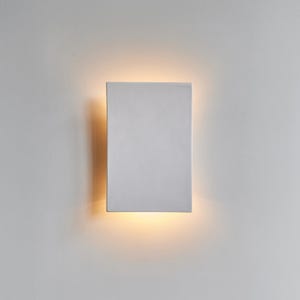 May include: A rectangular, white wall sconce with a warm, yellow glow emanating from the bottom. The sconce is mounted on a white wall, creating a minimalist and modern aesthetic. The light fixture is designed for indoor use.