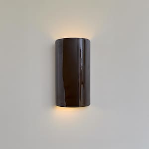 Könnte beinhalten: Zylindrische, dunkelbraune Wandleuchte mit glänzendem Finish. Die Leuchte strahlt ein warmes, nach unten gerichtetes Licht aus und erzeugt ein sanftes Leuchten. Die Leuchte ist an einer neutralfarbenen Wand montiert und bietet eine moderne Beleuchtungslösung.