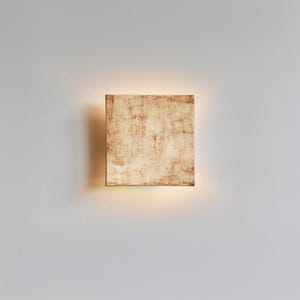 May include: Square wall light with a textured, cream and reddish-brown finish. The light is mounted on a white wall and emits a warm, diffused glow from the bottom. The design is minimalist and contemporary.