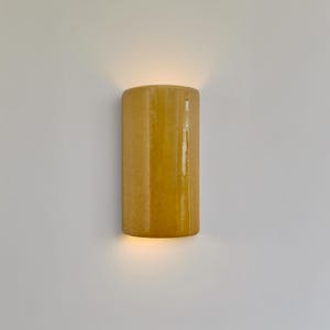 Op de afbeelding: Een cilindrische wandlamp met een warme gele kleur, die een zachte gloed uitstraalt. De armatuur is op een witte muur gemonteerd, wat een contrast creëert. Eenvoudig en modern ontwerp, geschikt voor binnenverlichting.