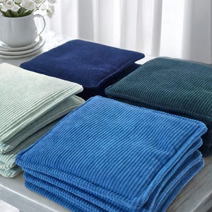 May include: A collection of four square corduroy cushions in shades of light green, dark blue, teal, and bright blue. Each cushion features a distinct ribbed texture and a square shape with stitched edges, stacked neatly in a group.