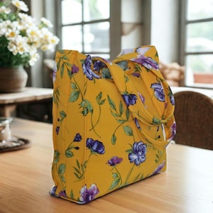 May include: A yellow tote bag featuring a floral design with purple and blue flowers and green foliage. The bag has two handles and is placed on a wooden table. The background shows a window and a vase of white flowers.