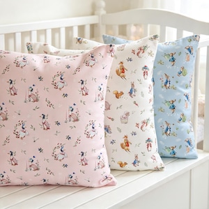 May include: Three square scatter cushions featuring repeating patterns of storybook characters. The left cushion is light pink with ducks, the centre is cream with woodland animals, and the right is light blue with rabbits in blue jackets.