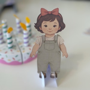 Pode incluir: Recorte de papel de uma menina de desenho animado com cabelo castanho e um la&ccedil;o vermelho. Ela usa uma camisa branca e macac&atilde;o cinza. Um bolo de anivers&aacute;rio com velas est&aacute; no fundo.