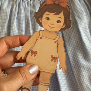 Pode incluir: Uma boneca de papel de uma menina com cabelo castanho e uma fita de cabelo com la&ccedil;o vermelho. Ela usa um macac&atilde;o bege com la&ccedil;os vermelhos e sapatos castanhos. A boneca &eacute; segurada contra um fundo xadrez azul e branco.