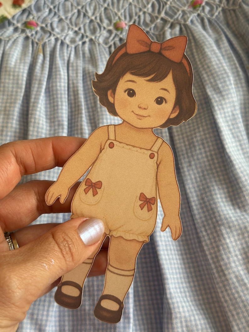 Pode incluir: Uma boneca de papel de uma menina com cabelo castanho e uma fita de cabelo com la&ccedil;o vermelho. Ela usa um macac&atilde;o bege com la&ccedil;os vermelhos e sapatos castanhos. A boneca &eacute; segurada contra um fundo xadrez azul e branco.