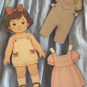 Pode incluir: Uma boneca de papel com um la&ccedil;o vermelho, vestindo um macac&atilde;o branco com la&ccedil;os vermelhos e sapatos marrons. Tamb&eacute;m est&atilde;o inclu&iacute;dos um macac&atilde;o bege e um vestido rosa com um desenho de cora&ccedil;&atilde;o. Em um fundo xadrez azul e branco.