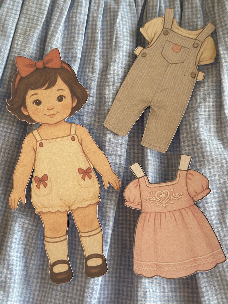 Pode incluir: Uma boneca de papel com um la&ccedil;o vermelho, vestindo um macac&atilde;o branco com la&ccedil;os vermelhos e sapatos marrons. Tamb&eacute;m est&atilde;o inclu&iacute;dos um macac&atilde;o bege e um vestido rosa com um desenho de cora&ccedil;&atilde;o. Em um fundo xadrez azul e branco.