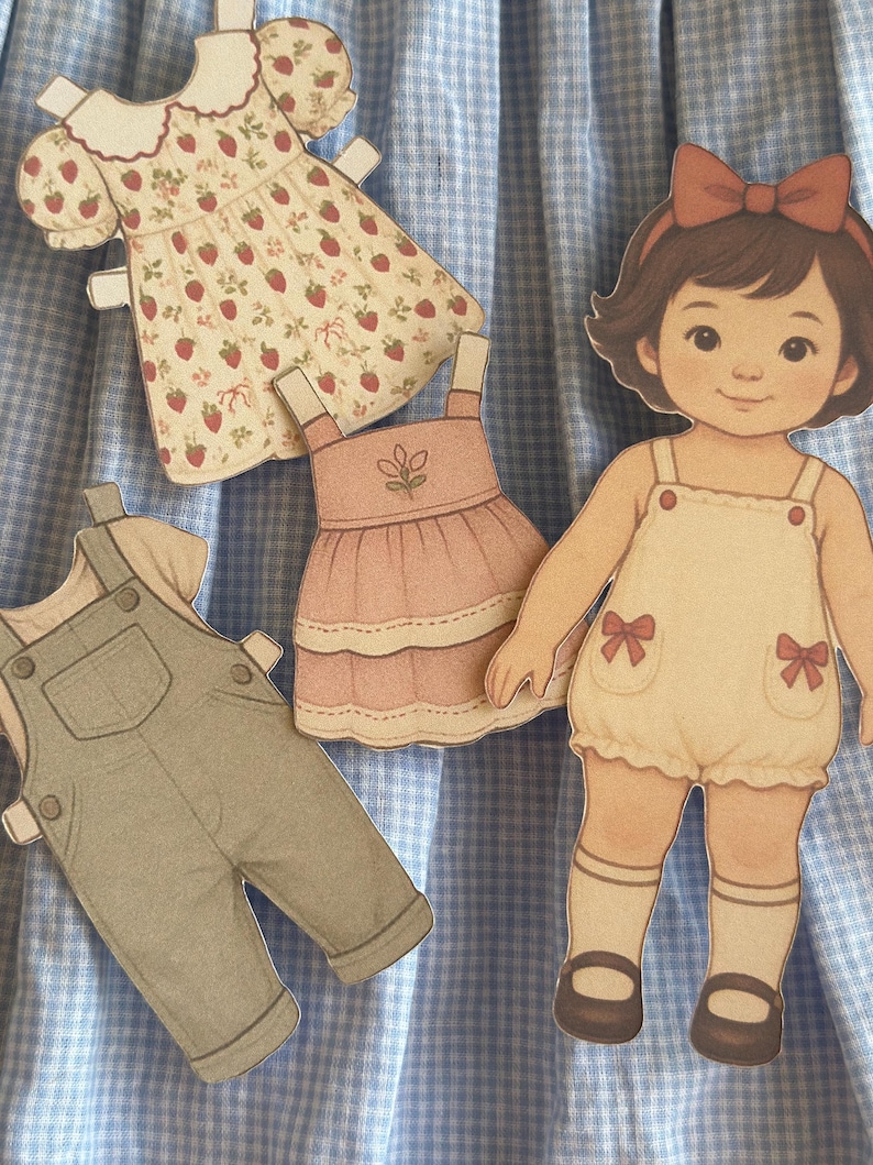 Pode incluir: Uma boneca de papel com um la&ccedil;o castanho e um macac&atilde;o creme com la&ccedil;os vermelhos. Existem tr&ecirc;s roupas de papel: um vestido com estampa de morango, um vestido rosa e um macac&atilde;o cinza.