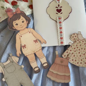 Pode incluir: Uma boneca de papel com um corte de cabelo castanho e um la&ccedil;o rosa, vestindo um macac&atilde;o creme com la&ccedil;os vermelhos. Roupas de papel adicionais incluem macac&otilde;es e vestidos. Um cart&atilde;o branco com um design em forma de cora&ccedil;&atilde;o e uma fita com detalhes de cora&ccedil;&atilde;o tamb&eacute;m est&aacute; presente.