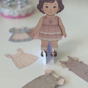 Pode incluir: Uma boneca de papel com um vestido rosa e um la&ccedil;o vermelho, com roupas de papel adicionais. A boneca tem cabelo castanho e est&aacute; em p&eacute; sobre um suporte branco. Roupas adicionais incluem um vestido e macac&atilde;o.