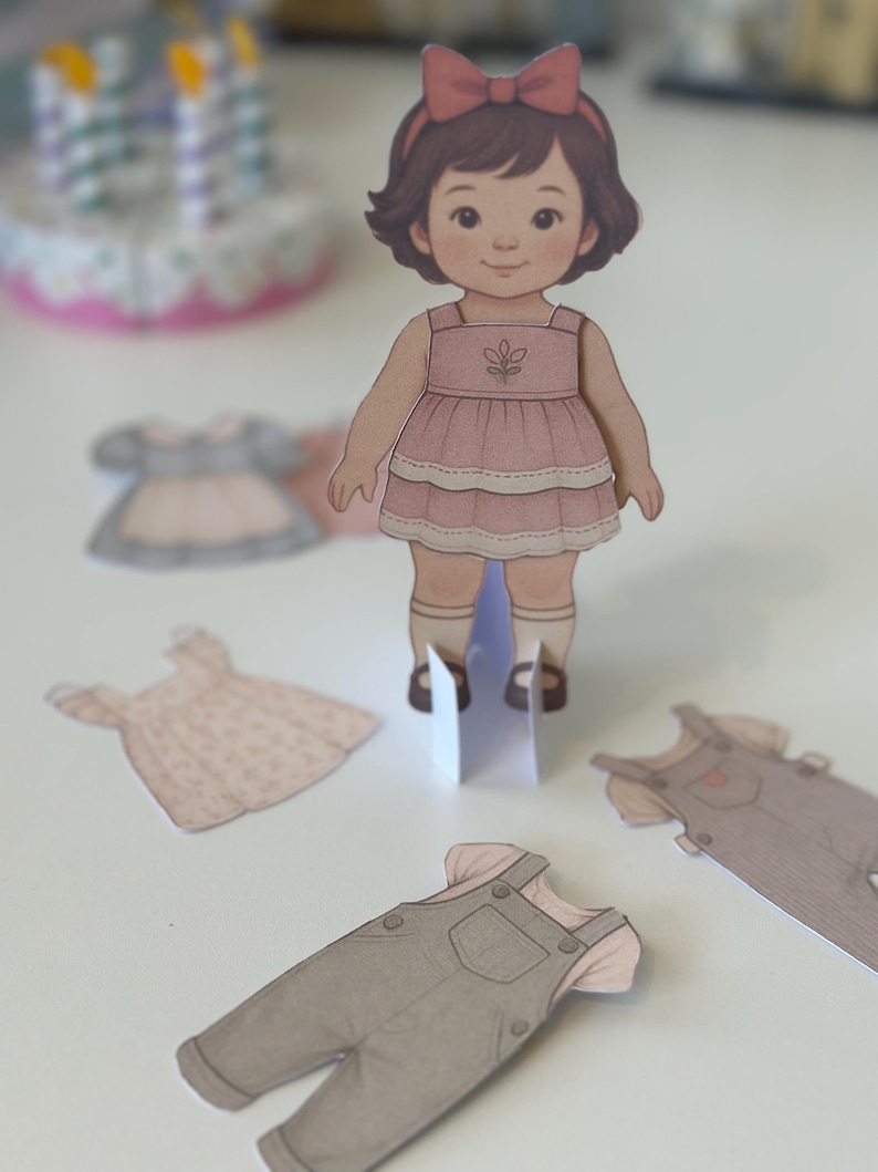 Pode incluir: Uma boneca de papel com um vestido rosa e um la&ccedil;o vermelho, com roupas de papel adicionais. A boneca tem cabelo castanho e est&aacute; em p&eacute; sobre um suporte branco. Roupas adicionais incluem um vestido e macac&atilde;o.