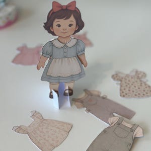 Pode incluir: Uma boneca de papel com cabelo castanho e um la&ccedil;o vermelho, vestindo um vestido azul com um avental branco. V&aacute;rias roupas de papel, incluindo vestidos e macac&otilde;es, est&atilde;o espalhadas ao redor da boneca em uma superf&iacute;cie branca.