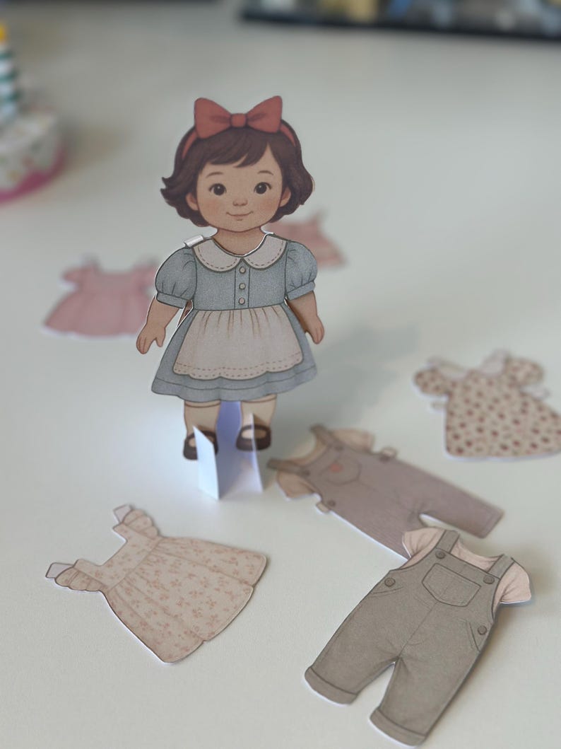 Pode incluir: Uma boneca de papel com cabelo castanho e um la&ccedil;o vermelho, vestindo um vestido azul com um avental branco. V&aacute;rias roupas de papel, incluindo vestidos e macac&otilde;es, est&atilde;o espalhadas ao redor da boneca em uma superf&iacute;cie branca.