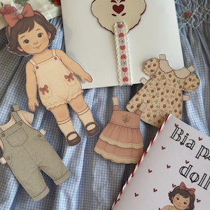 Pode incluir: Conjunto de boneca de papel com uma figura de boneca, macac&atilde;o e vestidos. A boneca tem um la&ccedil;o no cabelo e usa um romper. O conjunto inclui um livreto com o texto "Bia paper doll". O fundo &eacute; um padr&atilde;o xadrez azul e branco.