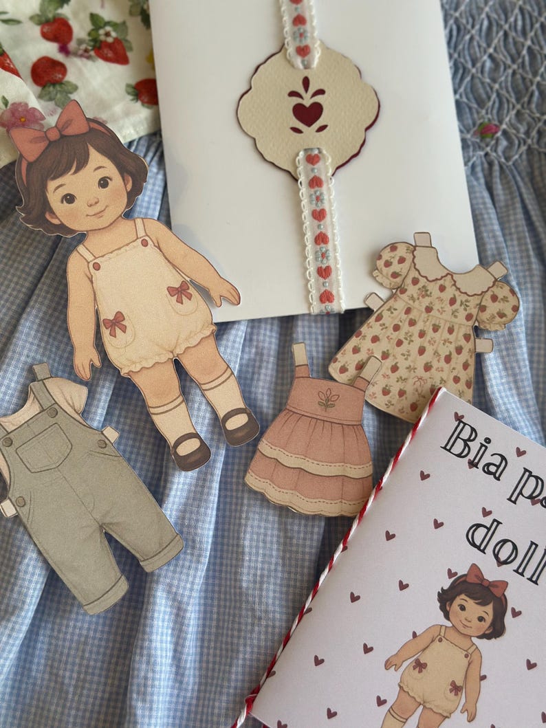 Pode incluir: Conjunto de boneca de papel com uma figura de boneca, macac&atilde;o e vestidos. A boneca tem um la&ccedil;o no cabelo e usa um romper. O conjunto inclui um livreto com o texto "Bia paper doll". O fundo &eacute; um padr&atilde;o xadrez azul e branco.