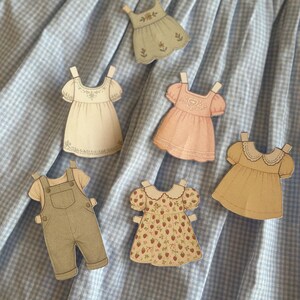 Pode incluir: Uma cole&ccedil;&atilde;o de recortes de roupas de bonecas de papel, incluindo vestidos e macac&otilde;es, em v&aacute;rias cores e padr&otilde;es. Os recortes s&atilde;o dispostos sobre um fundo de tecido xadrez azul e branco. Os vestidos apresentam detalhes como estampas florais e bordas de renda.