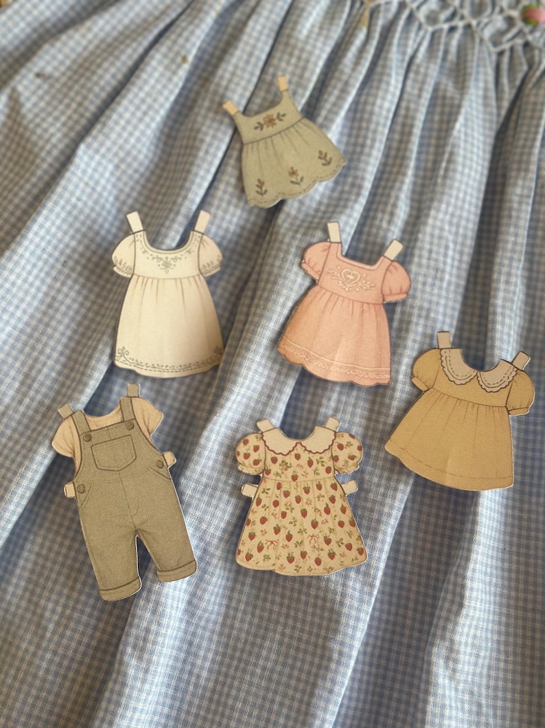 Pode incluir: Uma cole&ccedil;&atilde;o de recortes de roupas de bonecas de papel, incluindo vestidos e macac&otilde;es, em v&aacute;rias cores e padr&otilde;es. Os recortes s&atilde;o dispostos sobre um fundo de tecido xadrez azul e branco. Os vestidos apresentam detalhes como estampas florais e bordas de renda.