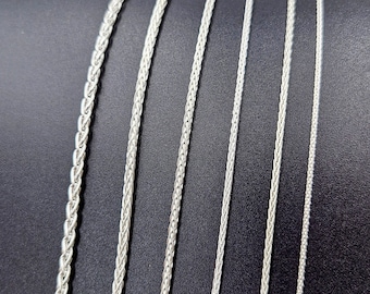 May include: A set of six silver chains of varying widths, displayed on a black surface. The chains are all made of a braided design.