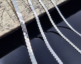 May include: Four silver chains of varying lengths, laid out on a textured surface. The chains are made of small, linked, flat, oval-shaped pieces.